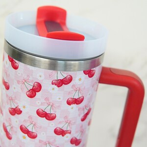 Puede incluir: Un termo de viaje de acero inoxidable con asa y tapa rojas. El termo presenta un estampado de cuadros rosas y blancos con ilustraciones de cerezas rojas y detalles de margaritas blancas. La tapa tiene un cierre abatible rojo.