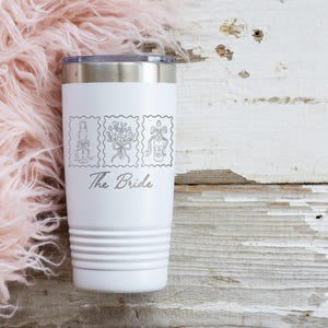 May include: A white stainless steel tumbler with a silver rim and a tapered base. The front features three black line-art illustrations of a champagne flute, a floral bouquet, and a bottle, with the text The Bride printed in a script font below.