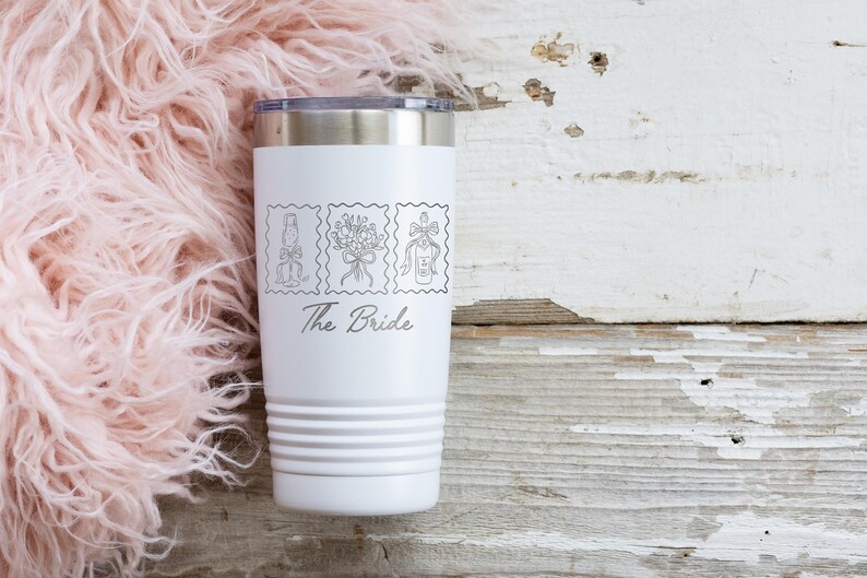 May include: A white stainless steel tumbler with a silver rim and a tapered base. The front features three black line-art illustrations of a champagne flute, a floral bouquet, and a bottle, with the text The Bride printed in a script font below.