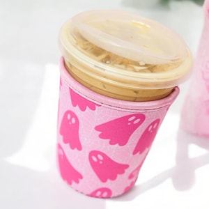 May include: A pink reusable coffee cup sleeve featuring a pattern of pink ghost illustrations. The cup contains a beverage and is topped with a clear plastic lid. The sleeve is designed to insulate the cup and protect the user's hands.