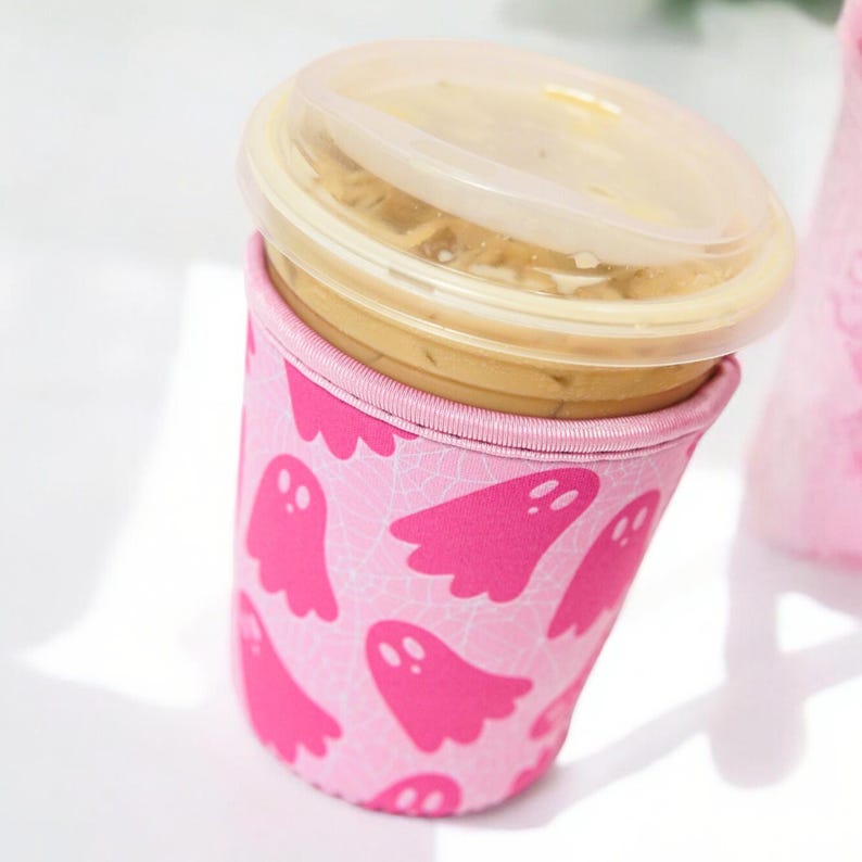 May include: A pink reusable coffee cup sleeve featuring a pattern of pink ghost illustrations. The cup contains a beverage and is topped with a clear plastic lid. The sleeve is designed to insulate the cup and protect the user's hands.