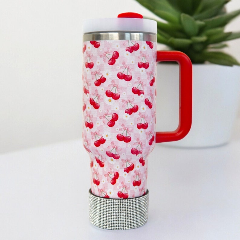 Puede incluir: Un termo de viaje de acero inoxidable con asa y tapa rojas. El termo est&aacute; decorado con un estampado de cuadros rosas y blancos con cerezas rojas y flores blancas. Una banda de diamantes de imitaci&oacute;n plateados adorna la base.