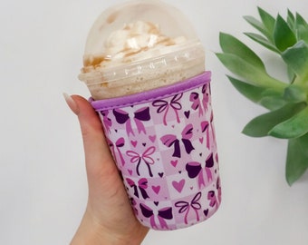 May include: A pink and purple insulated cup sleeve with a pattern of bows and hearts. The sleeve is on a clear plastic cup with a brown iced coffee drink and whipped cream.