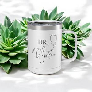 May include: White stainless steel travel mug with a black lid and handle. The mug has a black and white graphic of a stethoscope and the text "DR. Wilson".