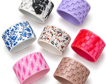 May include: Seven silicone rings featuring various designs: a black and white checkered pattern, a solid lavender checkerboard, a blue and white floral print, a multi-colored botanical pattern, a solid red checkerboard, a pink lightning bolt print, and a solid tan checkerboard.