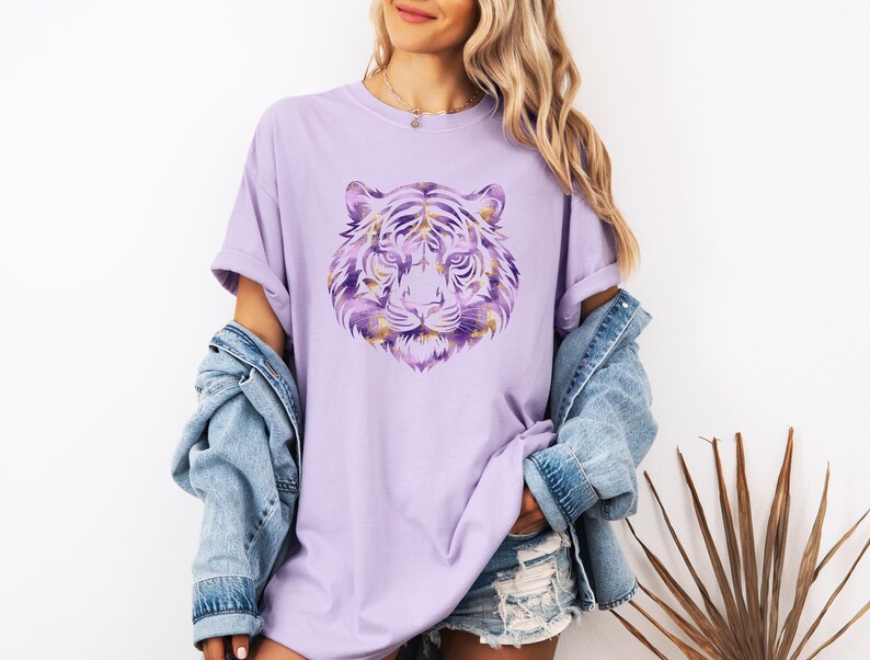 May include: A lavender t-shirt featuring a purple and gold tiger graphic. The tiger's face is centrally positioned on the shirt.