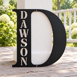 May include: A large, matte black metal wall sign shaped as the capital letter D. The vertical stem of the letter features the name DAWSON cut out in a bold, serif font. The sign includes two small circular mounting holes at the top and bottom.