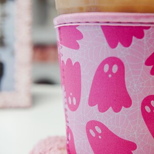 May include: A pink Halloween-themed drinks sleeve featuring a ghost and spiderweb design. The sleeve is around a clear plastic cup with a lid. The cup rests on a pink coaster, with a blurred pink frame in the background.