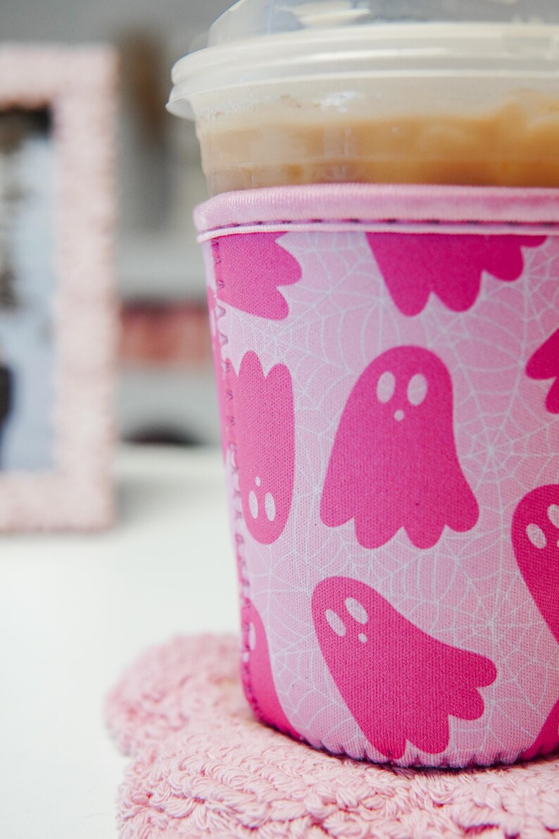 May include: A pink Halloween-themed drinks sleeve featuring a ghost and spiderweb design. The sleeve is around a clear plastic cup with a lid. The cup rests on a pink coaster, with a blurred pink frame in the background.