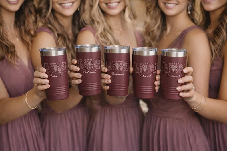 May include: Five maroon stainless steel tumblers with silver lids, each featuring a series of four white line-art illustrations of flowers and the word "Bridesmaid" engraved in a cursive script font on the front of the cylindrical vessel.