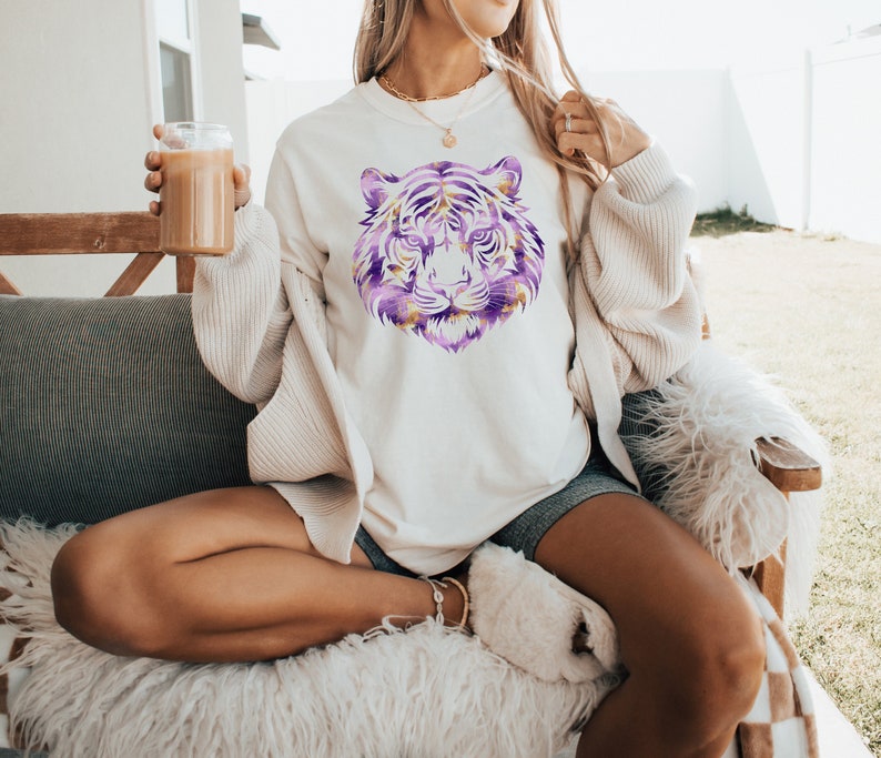 May include: A white sweatshirt featuring a purple and gold tiger face graphic. The model is wearing the sweatshirt with a white cardigan and sitting on a fluffy white chair.