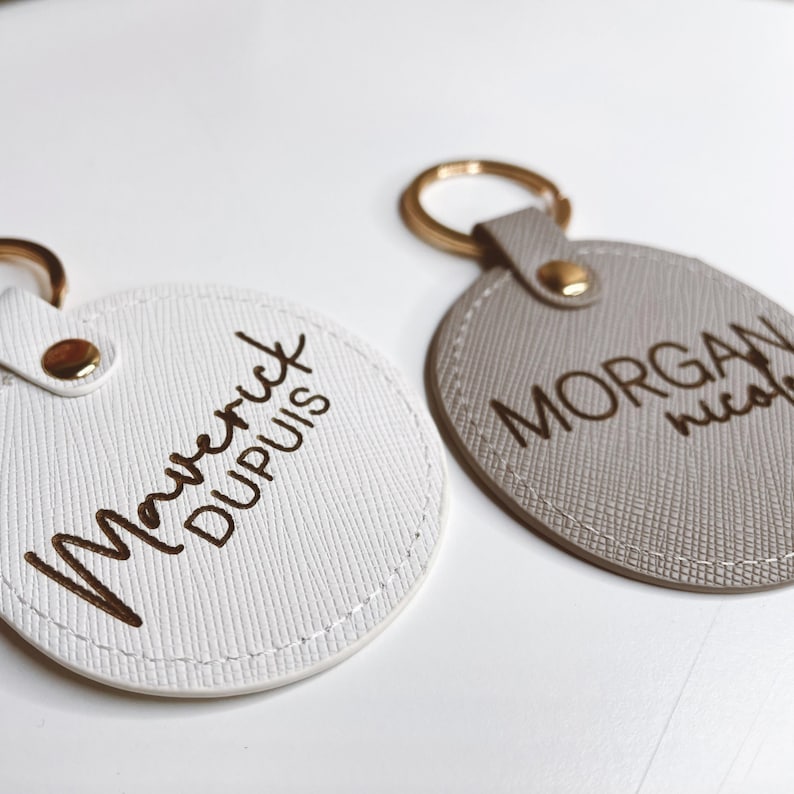 Pu&ograve; includere: Due portachiavi personalizzati. Uno &egrave; bianco con "Maverick Dupuis" in lettere dorate. L'altro &egrave; grigio con "Morgan Nicole" in lettere dorate. Entrambi hanno portachiavi dorati e rivetti dorati abbinati.