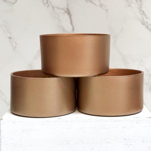 May include: Three stacked, round, metallic gold-colored planters. The planters are made of a smooth, matte material.