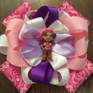 May include: A pink, white, and purple hair bow with a cartoon pirate girl in the center. The bow is made of ribbon and has a paisley pattern.