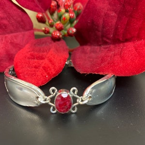 May include: A silver bracelet with a red gemstone set in a silver filigree design. The bracelet is made from two spoon halves.
