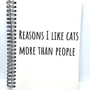 May include: A white notebook with a black spiral binding, featuring the text "REASONS I LIKE CATS MORE THAN PEOPLE" in black. The text is centered on the page, making a statement about cat preference.