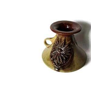 May include: A small ceramic vessel with a wide, flared base and a narrow neck featuring a dark brown glazed rim. The body is a muted tan colour with a prominent, raised floral relief pattern in dark brown and a single small side handle.