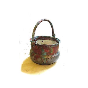 May include: A small, antique copper cauldron with a twisted handle. The cauldron has a rounded base and shows signs of age with a weathered, reddish-brown and turquoise patina. The handle is a darker, twisted rope design.