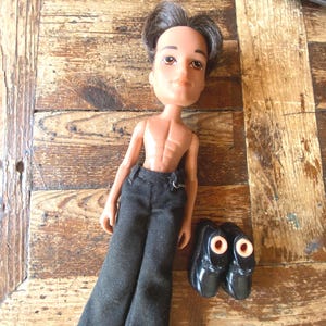 Könnte beinhalten: Eine Bratz-Puppe mit dunklem Haar, gebräunter Haut und schwarzer Hose. Die Puppe ist ohne Hemd und hat einen muskulösen Körperbau. Ein Paar schwarze Schuhe mit orangefarbenen Akzenten befindet sich neben der Puppe. Die Puppe befindet sich auf einer Holzoberfläche.