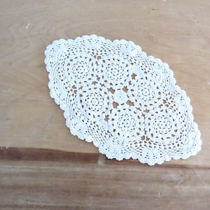 May include: Cream-colored, oval-shaped crochet doily with a scalloped edge. The doily features a repeating floral pattern in the center and along the border. The handmade textile is displayed on a light brown wooden surface.