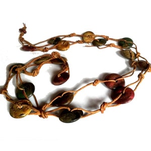 May include: A necklace featuring multiple round, flat, multi-colored wooden discs in shades of red, green, and brown. The discs are strung along a thin, light brown cord that is knotted at intervals to secure each piece in a fixed position.