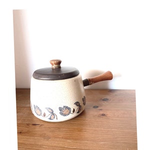 May include: Cream-colored ceramic saucepan with a dark brown lid and wooden handle. The saucepan features a floral pattern in blue and brown. The handle is attached to the side of the pan. The saucepan is sitting on a wooden surface.