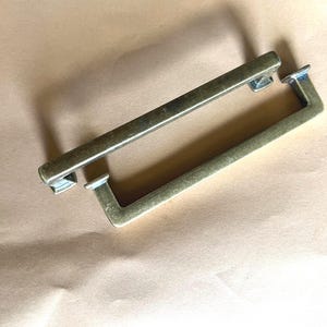 Puede incluir: Dos tiradores de cajón rectangulares de metal con un acabado de latón antiguo cepillado. Cada tirador presenta una barra larga y recta con dos postes de montaje perpendiculares en los extremos, creando un diseño simple y funcional.