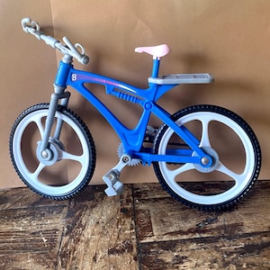 Op de afbeelding: Een blauw en witte speelgoedfiets met een roze zadel. De fiets heeft witte wielen met een blauw frame en stuur. De fiets staat op een houten oppervlak.