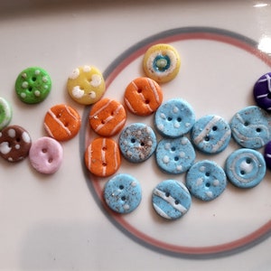 May include: A collection of round, two-hole buttons in various colors including green, yellow, orange, blue, purple, brown, and pink. The buttons feature diverse surface patterns such as white dots, stripes, swirls, and glittery textures.