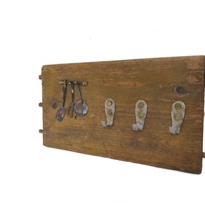 Puede incluir: Un perchero de pared rústico de madera con cuatro ganchos metálicos y una fila de utensilios de cocina en miniatura. La madera tiene un aspecto envejecido y los ganchos y utensilios metálicos muestran signos de oxidación.