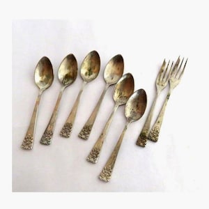 Puede incluir: Un conjunto de cubiertos antiguos que incluye seis cucharas y dos tenedores. Las cucharas tienen cuencos ovalados y detalles florales ornamentados en los mangos. La cubertería presenta un aspecto envejecido y deslustrado.
