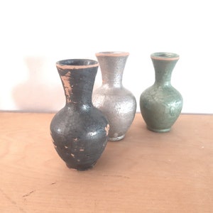 Puede incluir: Tres pequeños jarrones de cerámica en diferentes colores. El jarrón de la izquierda es gris oscuro, el jarrón del medio es plateado y el jarrón de la derecha es verde.