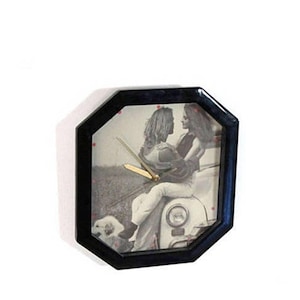 Puede incluir: Reloj octogonal negro con una imagen en blanco y negro de dos personas abrazándose en un vehículo vintage. El reloj tiene manecillas doradas y puntos rojos que marcan las horas. Un perro también está en la imagen.