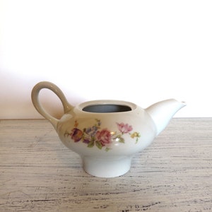 May include: A white ceramic creamer with a curved handle and spout. The creamer features a floral design in shades of pink, purple, and blue. The creamer is approximately 6 inches long and 4 inches tall.