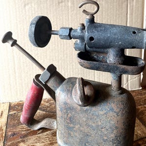 May include: A vintage metal blowtorch featuring a heavily rusted dark gray finish, a red wooden handle, a pump plunger, and a circular adjustment knob. The tool includes a burner nozzle assembly with a small metal loop at the top and a drip tray.