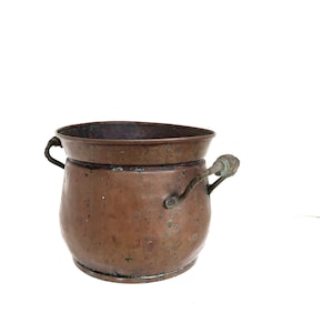 Pode incluir: Um pote de cobre redondo com uma borda larga e alargada, apresentando uma pátina escura e desgastada. O recipiente possui duas alças laterais de metal fixadas com rebites, exibindo sinais de oxidação e desgaste na superfície externa e interna.