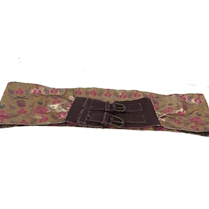 Puede incluir: Cinturón ancho y decorativo con un estampado floral en tonos dorados, rosas y marrones. El cinturón presenta un panel marrón oscuro con dos hebillas metálicas. El cinturón está diseñado para usarse alrededor de la cintura.