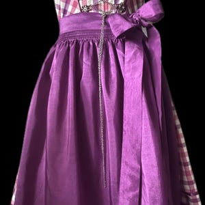 May include: A traditional dirndl dress with a purple skirt and a checkered bodice in shades of pink and white. The bodice features decorative metal clasps and a silver chain. A matching purple bow adorns the waist.