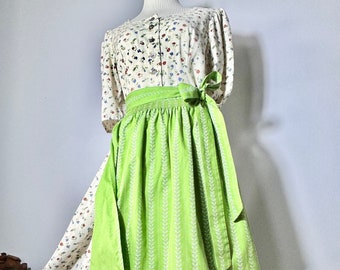 May include: A white and green dirndl dress with a floral print bodice and a green apron with a white leaf pattern. The apron has a green ribbon tied at the waist.