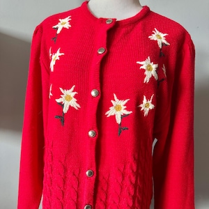 May include: A bright red cardigan sweater with embroidered white and yellow flower details. The sweater has a round neckline, long sleeves, and silver buttons. The lower front features a textured cable knit design.