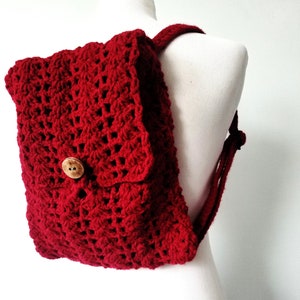 May include: A red crocheted backpack with a wooden button. The backpack is on a white mannequin.