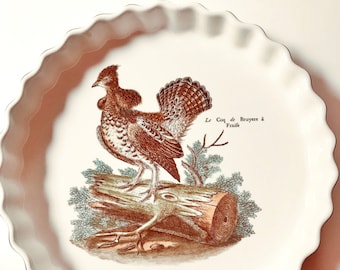 Könnte beinhalten: Eine weiße Keramik-Backform mit gewelltem Rand. Die Form zeigt eine braune und weiße Illustration eines Auerhuhns, das auf einem Baumstamm sitzt, mit dem Text "Le Coq de Bruyere a Fraide".