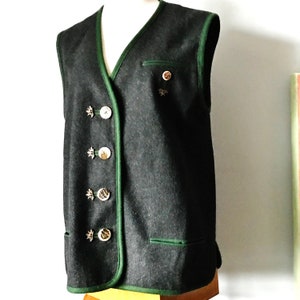 Puede incluir: Un chaleco verde oscuro con ribete verde y botones plateados. El chaleco tiene un bolsillo en el lado izquierdo y un broche decorativo en el lado derecho.
