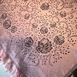 Puede incluir: Textil rosa con un estampado floral con rosas y hojas en un tono más oscuro. La tela tiene flecos en el borde. El diseño es un patrón repetitivo de flores y vides.