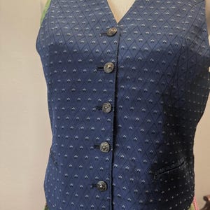 May include: A navy blue vest with a diamond pattern and silver-colored buttons. The vest has a V-neck and a small pocket on the right side. The vest is part of a traditional outfit.
