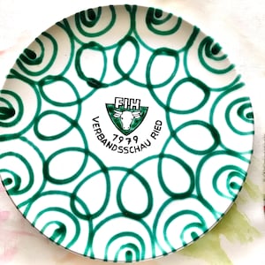 May include: White ceramic plate with a green and white swirl pattern. The center of the plate features a black and white logo with the text "FIH VERBANDSSCHAU 1979 RIED".