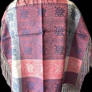 May include: A rectangular shawl featuring a patchwork design of floral patterns in shades of red, blue, and cream. The fabric displays a woven texture with repeating rose motifs across alternating colored squares, finished with long, thin fringe along the edges.
