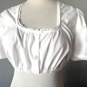 Puede incluir: Una blusa blanca de manga corta, con cuello cuadrado y tres botones en la parte delantera. El cuello y los bordes de las mangas están adornados con encaje. La cintura está fruncida. La blusa es de tejido ligero.