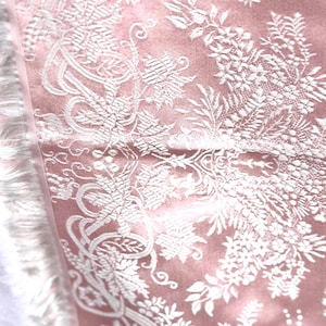 May include: Close-up of a dusty rose textile with intricate white floral embroidery. The design features detailed leaves, vines, and swirling patterns. The edge has a white fringe, adding texture and detail to the fabric.