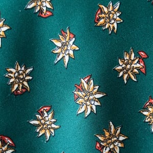 May include: A deep emerald green fabric featuring a repeating pattern of edelweiss flowers. Each flower is rendered in shades of white, yellow, and brown, with small red accents on the petals, creating a consistent floral motif across the surface.